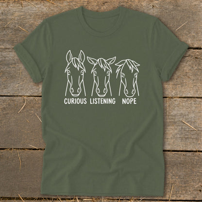 An olive green t-shirt featuring three horse head outline graphics with captions curious, listening, and nope.