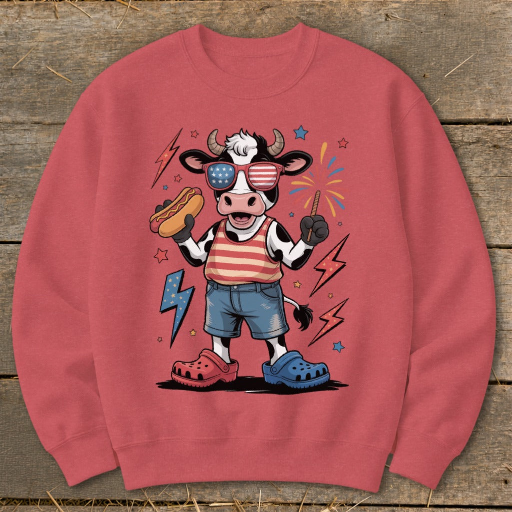 Cartoon cow in American flag sunglasses holding hot dog and sparkler.