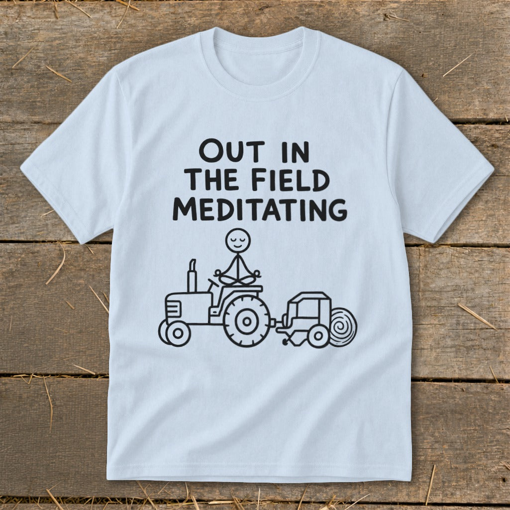 Light blue t-shirt with black text "OUT IN THE FIELD MEDITATING" and stick figure on tractor.