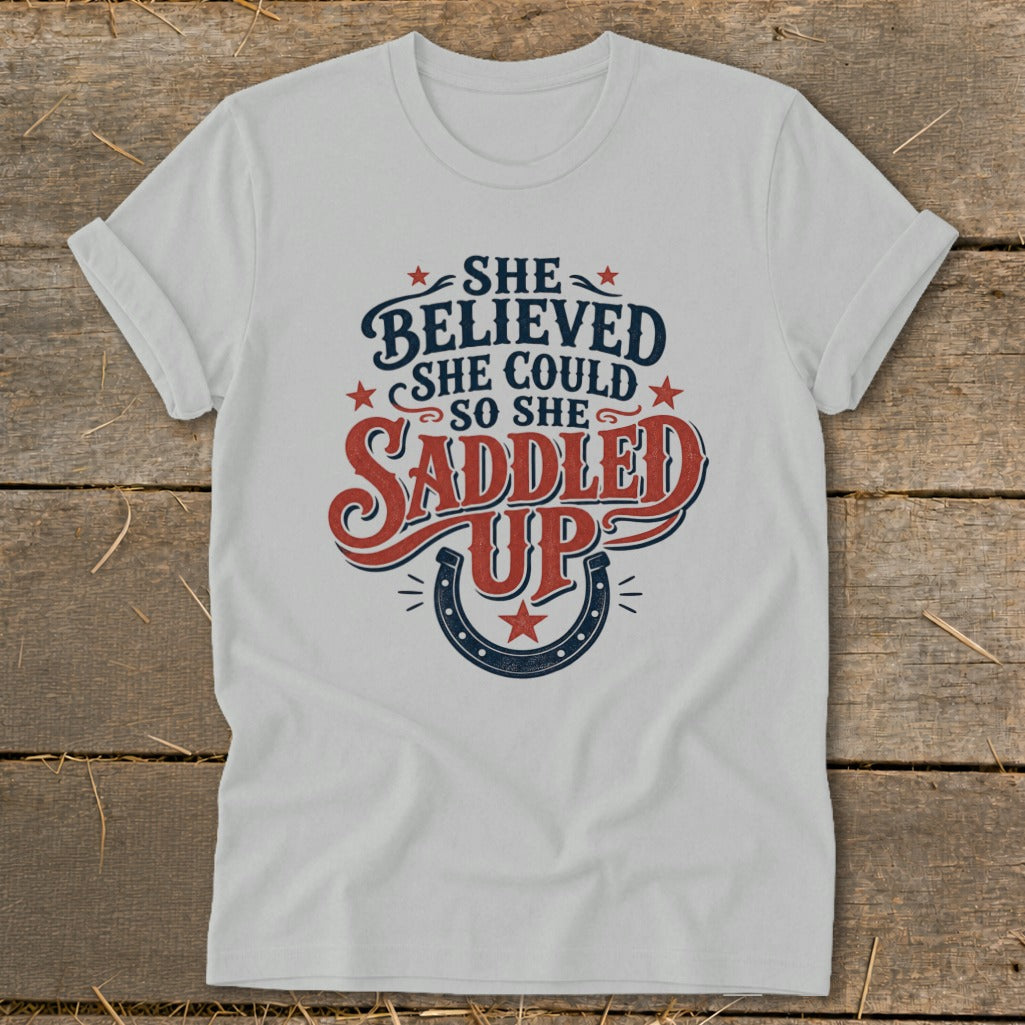 White t-shirt with "She believed she could so she saddled up" text and horseshoe graphic.