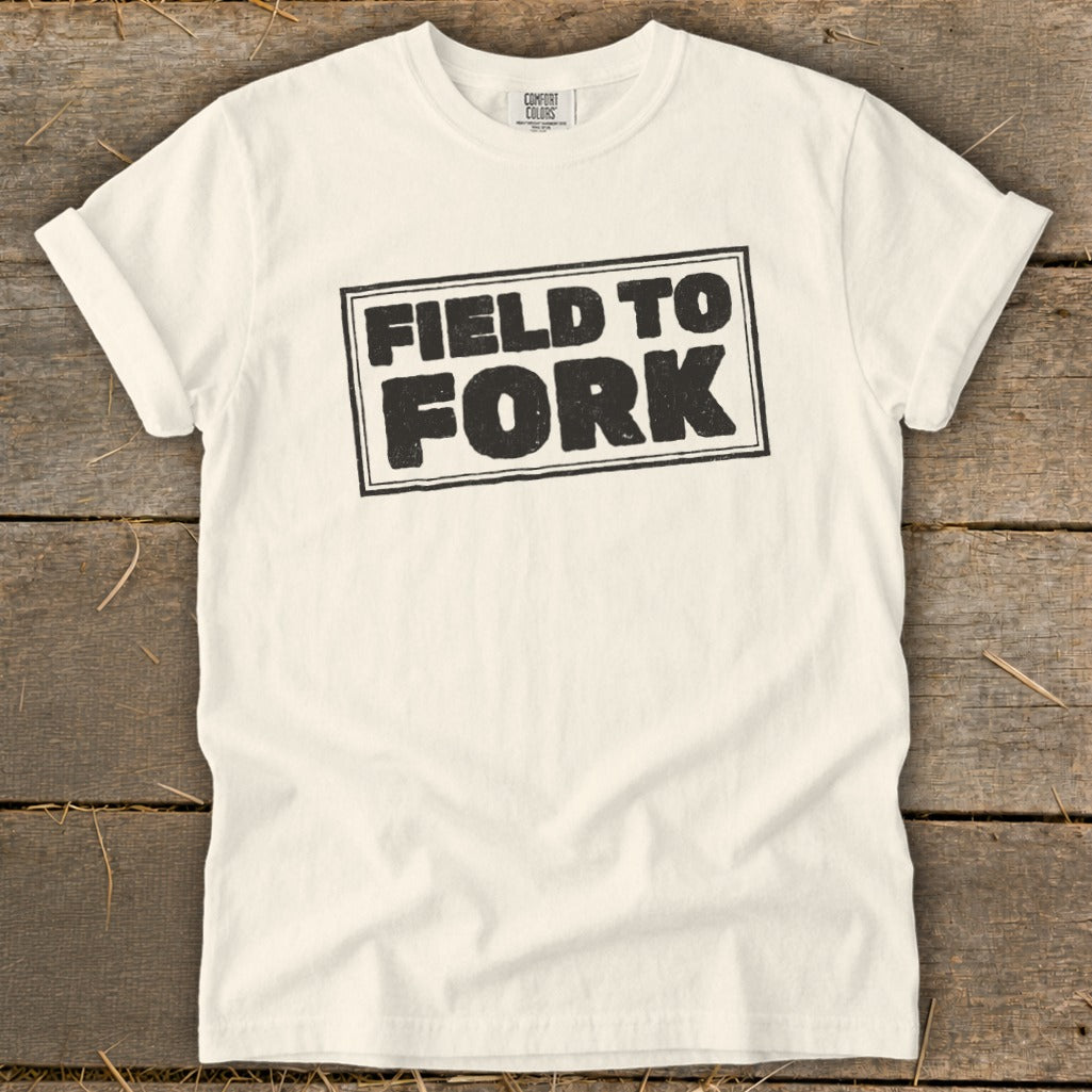A cream-colored t-shirt with rolled sleeves lies flat on a wooden surface, featuring black text that reads "FIELD TO FORK" inside a bordered rectangle.