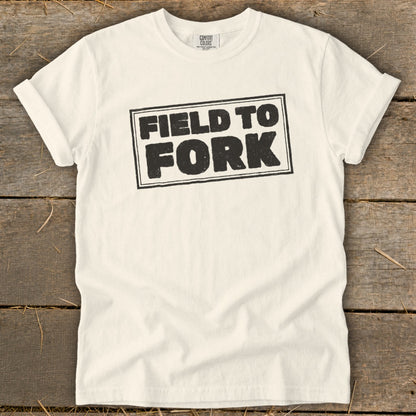 A cream-colored t-shirt with rolled sleeves lies flat on a wooden surface, featuring black text that reads "FIELD TO FORK" inside a bordered rectangle.