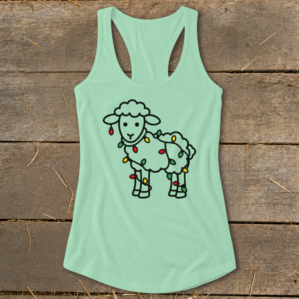 A light green women's racerback tank top featuring a black outline of a sheep wrapped in Christmas lights, laid flat on a wooden surface.