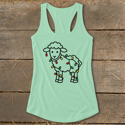 A light green women's racerback tank top featuring a black outline of a sheep wrapped in Christmas lights, laid flat on a wooden surface.