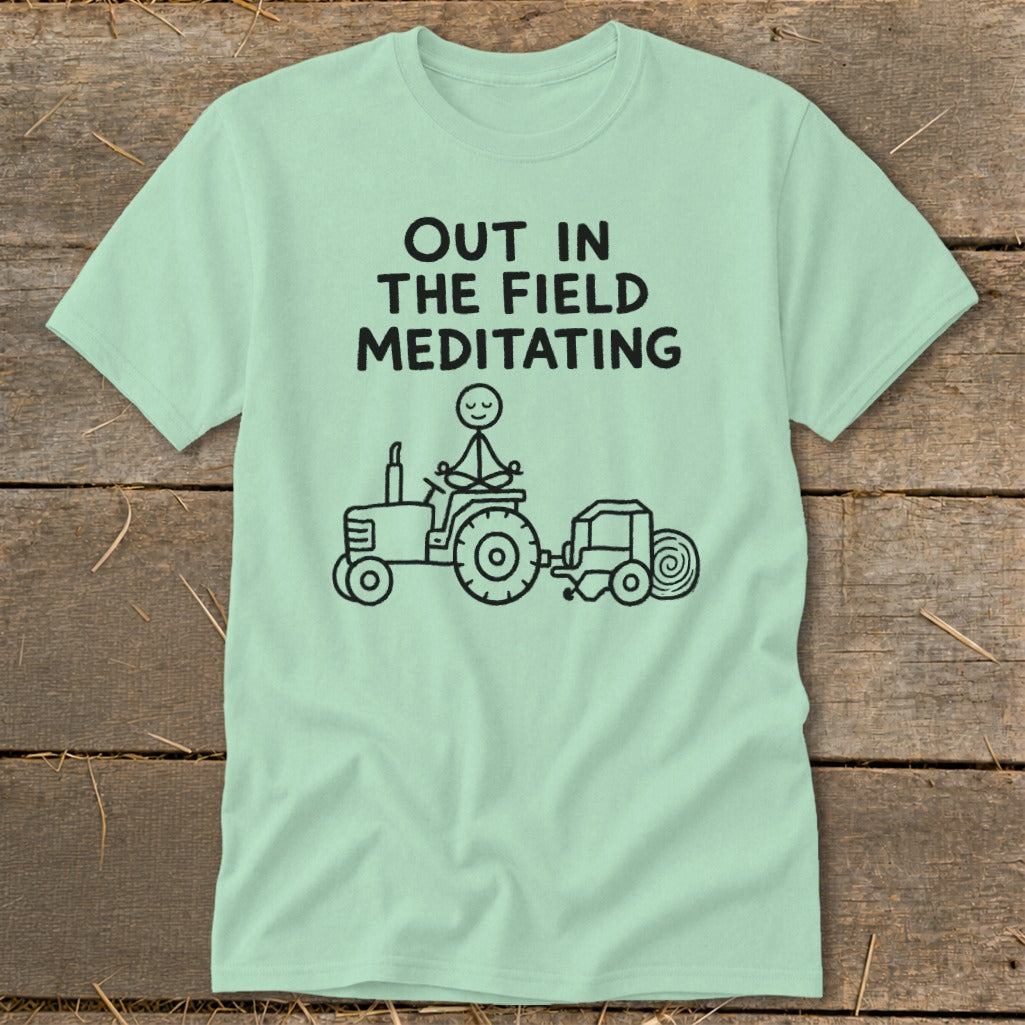 Light blue t-shirt with black text "OUT IN THE FIELD MEDITATING" and a stick figure meditating on a tractor.