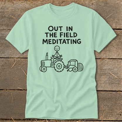 Light blue t-shirt with black text "OUT IN THE FIELD MEDITATING" and a stick figure meditating on a tractor.