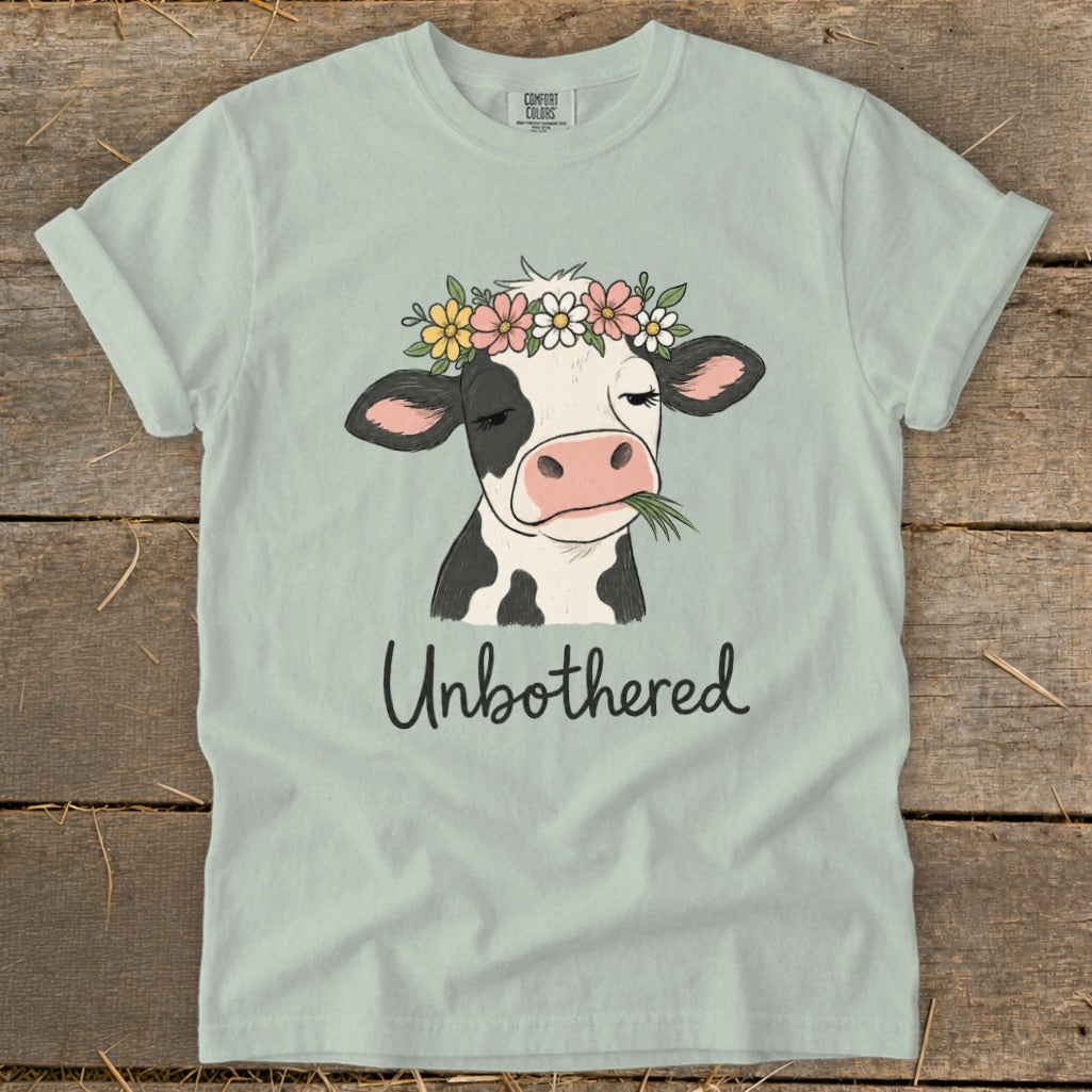 Light blue t-shirt with a cow wearing a floral crown and eating grass, text below reads Unbothered.