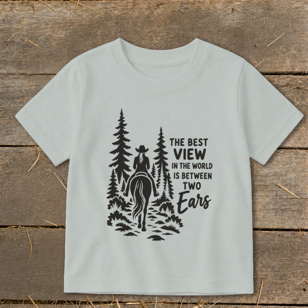 A light gray children's t-shirt features a black graphic of a horse and rider surrounded by trees and the text The Best View in the World Is Between Two Ears.