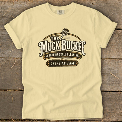 Pale yellow t-shirt with "The Muck Bucket School of Stall Cleaning Opens at 5 AM" graphic.