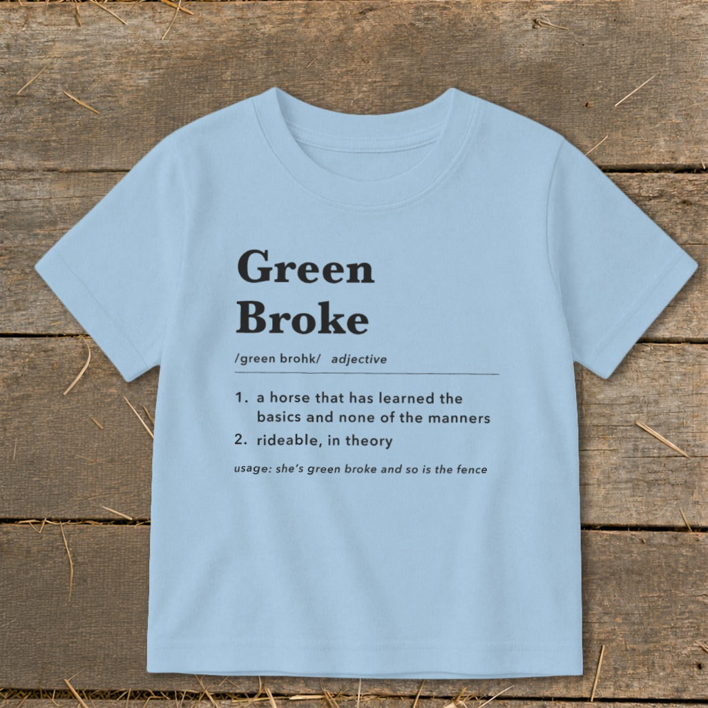 A light blue t-shirt printed with a dictionary definition of Green Broke on a rustic wooden background.