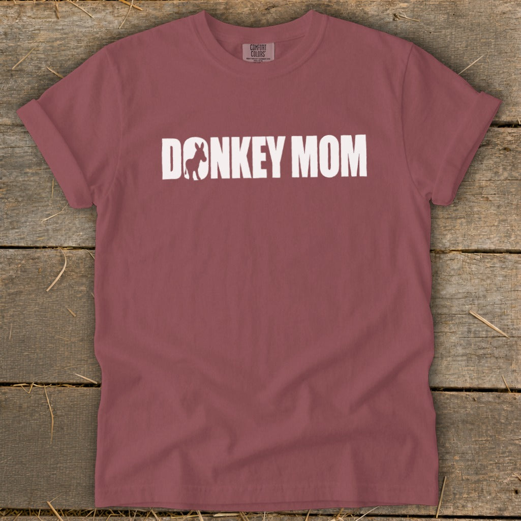A mauve short-sleeve t-shirt with white text reading DONKEY MOM on a wooden background.