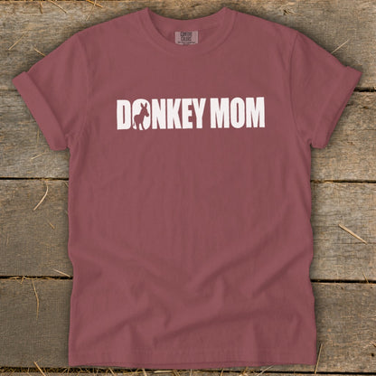 A mauve short-sleeve t-shirt with white text reading DONKEY MOM on a wooden background.