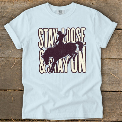 Light blue t-shirt with distressed graphic of a bronc rider and text "STAY LOOSE & STAY ON".