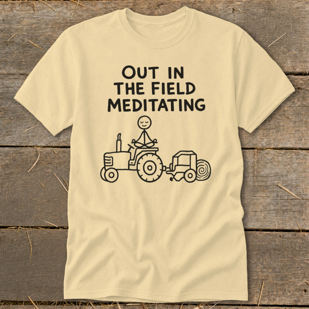Light yellow t-shirt with black text "OUT IN THE FIELD MEDITATING" and stick figure on tractor.