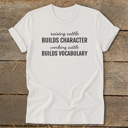 White t-shirt with black text reading "raising cattle BUILDS CHARACTER working cattle BUILDS VOCABULARY."