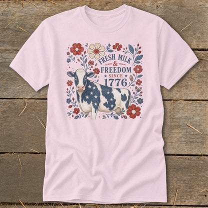 Pink t-shirt with a cow decorated in stars and stripes, surrounded by flowers, with text "Fresh Milk & Freedom Since 1776".