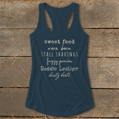 Navy tank top with white text listing equestrian items: sweet feed, warm barn, stall shavings, fuzzy ponies, saddle leather, dusty boots.
