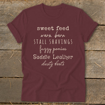 Maroon t-shirt with white text listing: sweet feed, warm barn, stall shavings, fuzzy ponies, saddle leather, dusty boots.