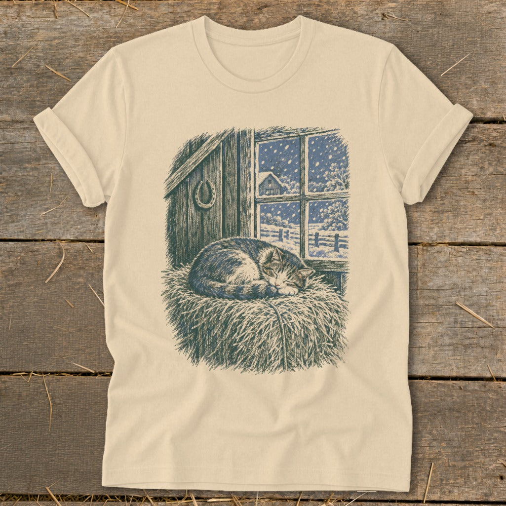 T-shirt featuring a sleeping cat on hay with a snowy farm scene outside a window.