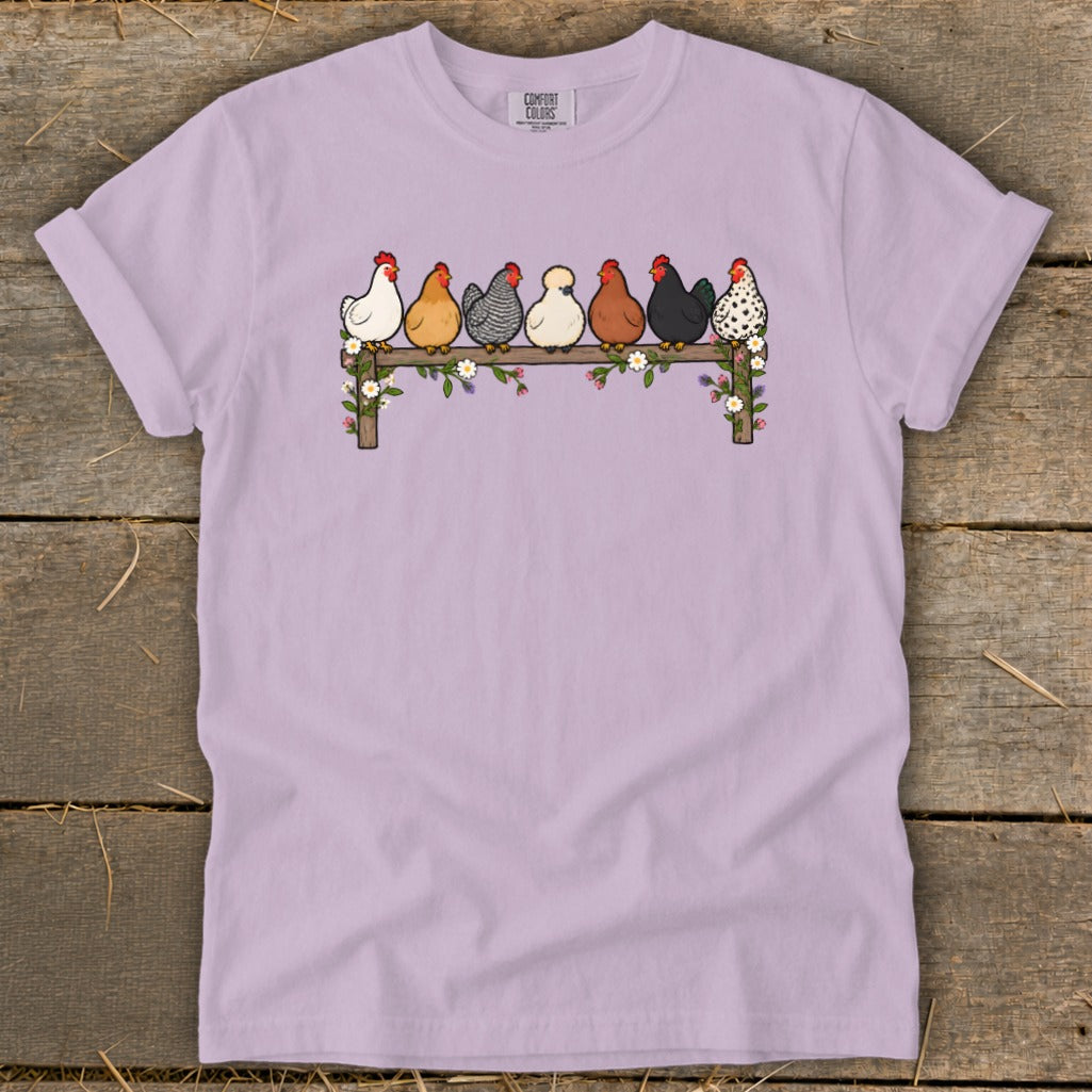 Light purple t-shirt featuring a graphic of seven colorful chickens perched on a flower-covered wooden fence.
