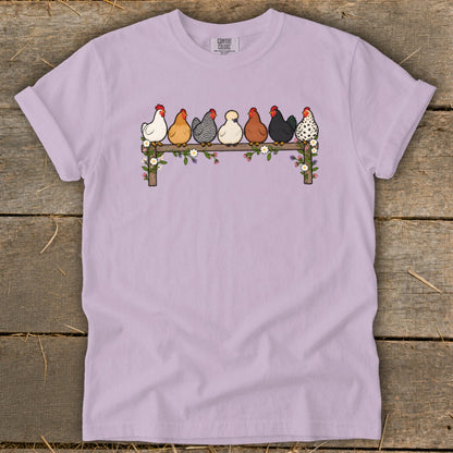 Light purple t-shirt featuring a graphic of seven colorful chickens perched on a flower-covered wooden fence.