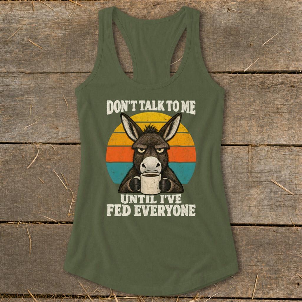 An olive green racerback tank top featuring a graphic of a grumpy donkey holding a coffee mug in front of a retro sunset and the text DON'T TALK TO ME UNTIL I'VE FED EVERYONE.