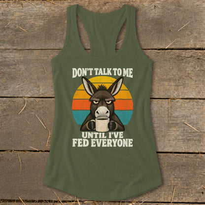An olive green racerback tank top featuring a graphic of a grumpy donkey holding a coffee mug in front of a retro sunset and the text DON'T TALK TO ME UNTIL I'VE FED EVERYONE.