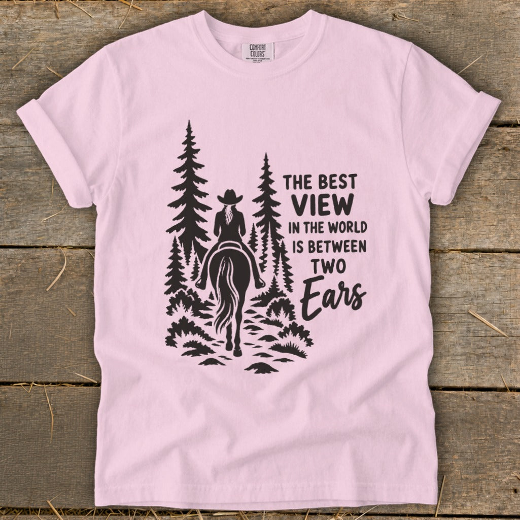A light pink t-shirt featuring a black graphic of a horseback rider in the woods and text reading, "The best view in the world is between two ears."