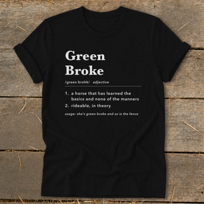 Black t-shirt featuring dictionary-style text defining the term green broke laid flat on a rustic wooden surface.