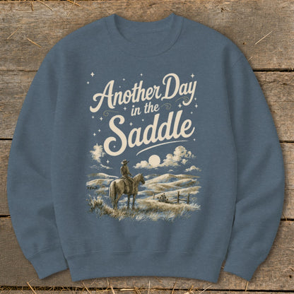 Crewneck sweatshirt with "Another Day in the Saddle" text over cowboy on horseback in a landscape.
