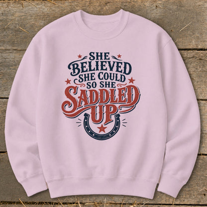 Pink sweatshirt with "She Believed She Could So She Saddled Up" text and horseshoe graphic.