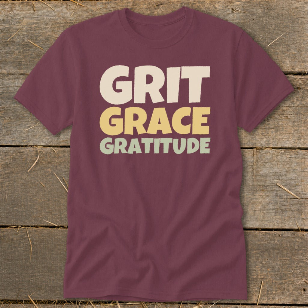 Maroon t-shirt with block lettering that reads "GRIT GRACE GRATITUDE" laid flat on wooden planks.