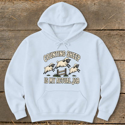 Light blue hoodie with a graphic of sheep jumping over a fence and text that reads "COUNTING SHEEP IS MY ACTUAL JOB".