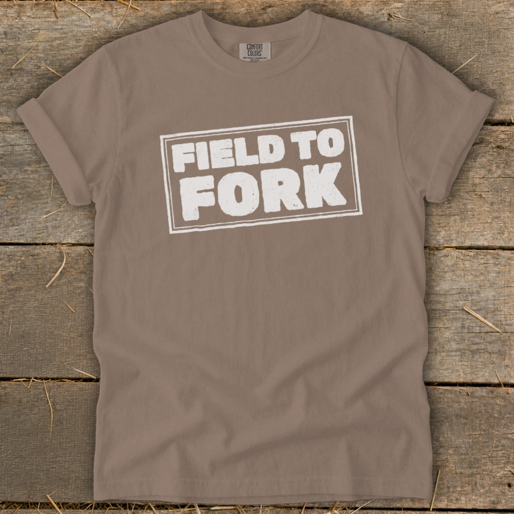Brown t-shirt featuring a white rectangular graphic with the text Field to Fork, laid flat on a wooden background.