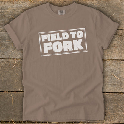 Brown t-shirt featuring a white rectangular graphic with the text Field to Fork, laid flat on a wooden background.