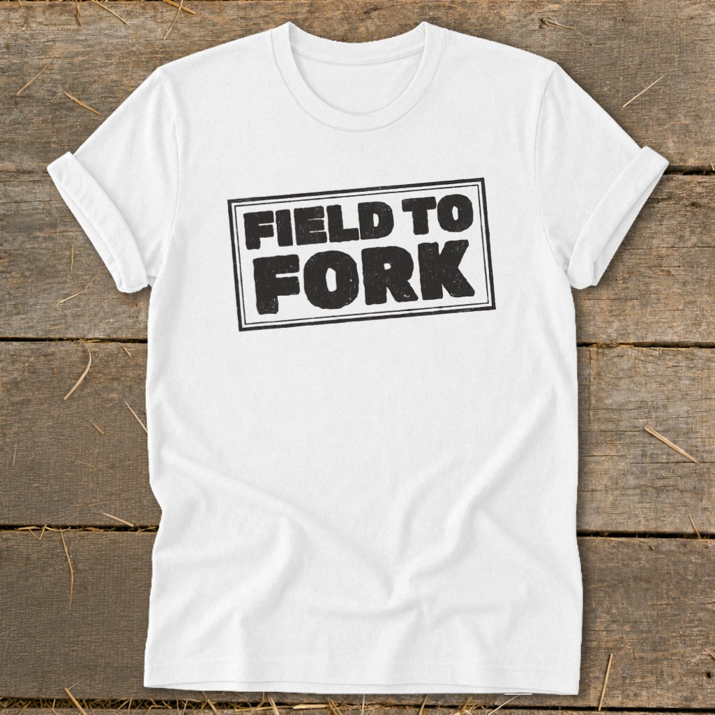 White t-shirt featuring the black text "FIELD TO FORK" inside a rectangular border, laid flat on a wooden surface.
