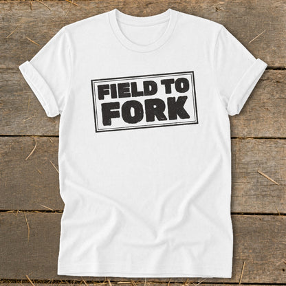 White t-shirt featuring the black text "FIELD TO FORK" inside a rectangular border, laid flat on a wooden surface.