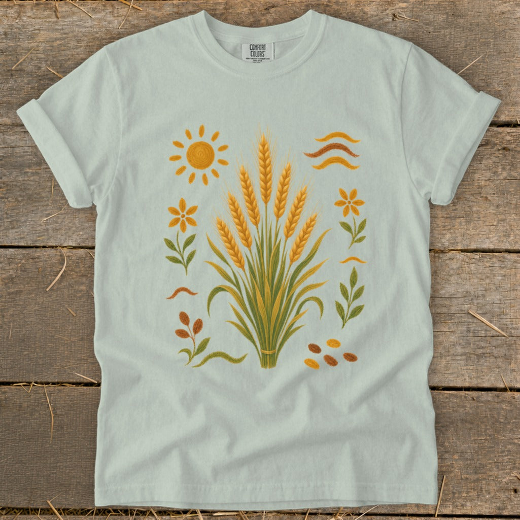 Light blue t-shirt with wheat stalks, sun, flowers, leaves, and seeds graphic.