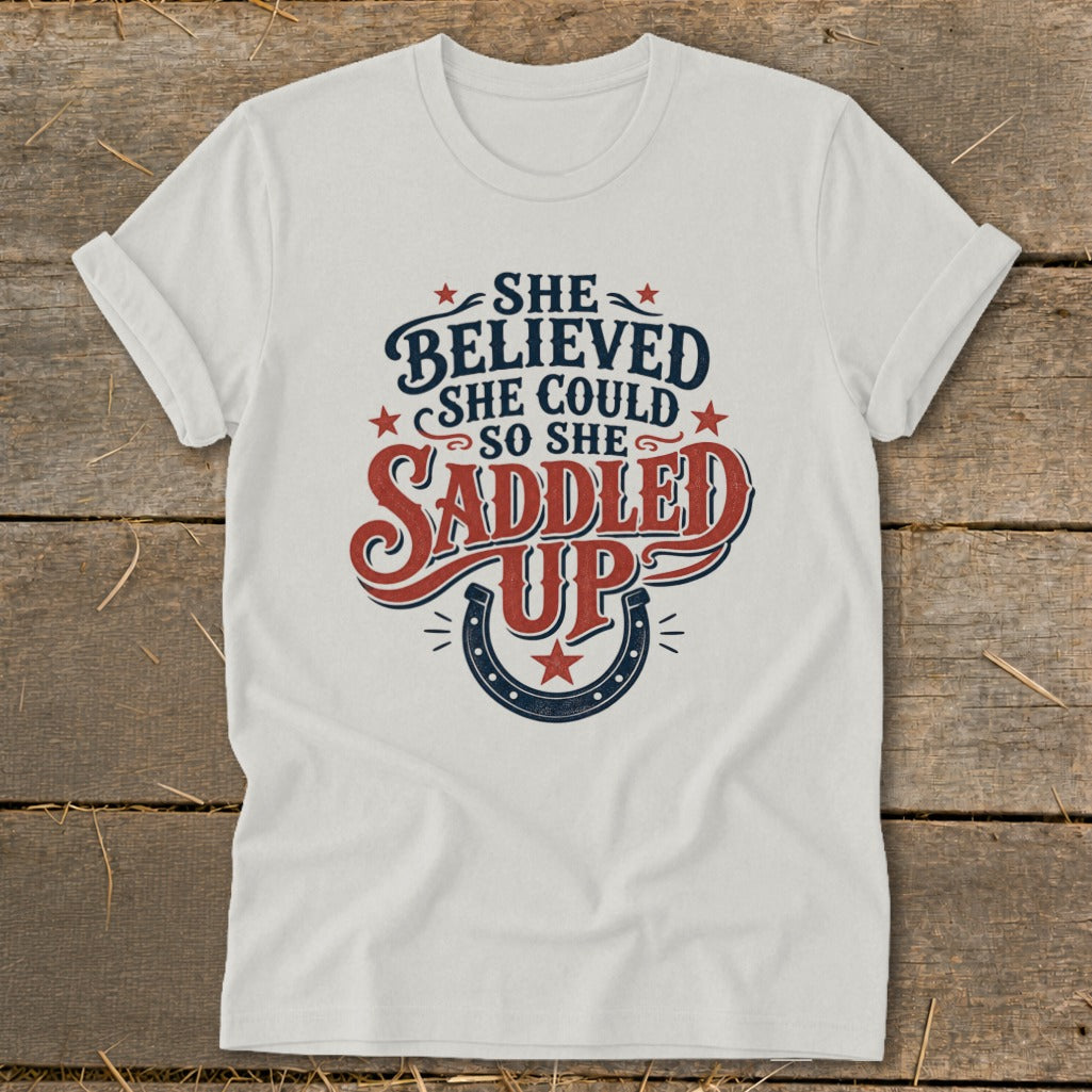 White t-shirt with "She Believed She Could So She Saddled Up" text and horseshoe graphic.