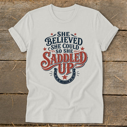 White t-shirt with "She Believed She Could So She Saddled Up" text and horseshoe graphic.