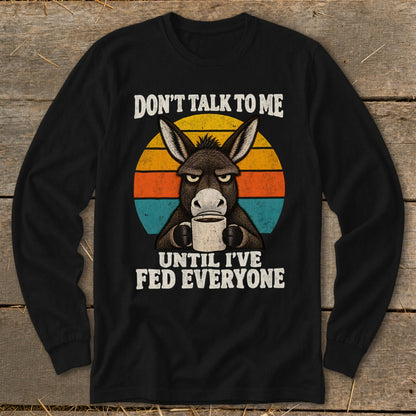 A black long-sleeved shirt with a graphic of an annoyed donkey holding a coffee mug against a retro sunset.