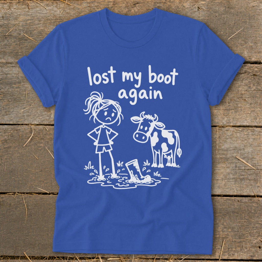 A blue t-shirt printed with a white graphic of a sad girl and a cow looking at a muddy boot, accompanied by the text lost my boot again.