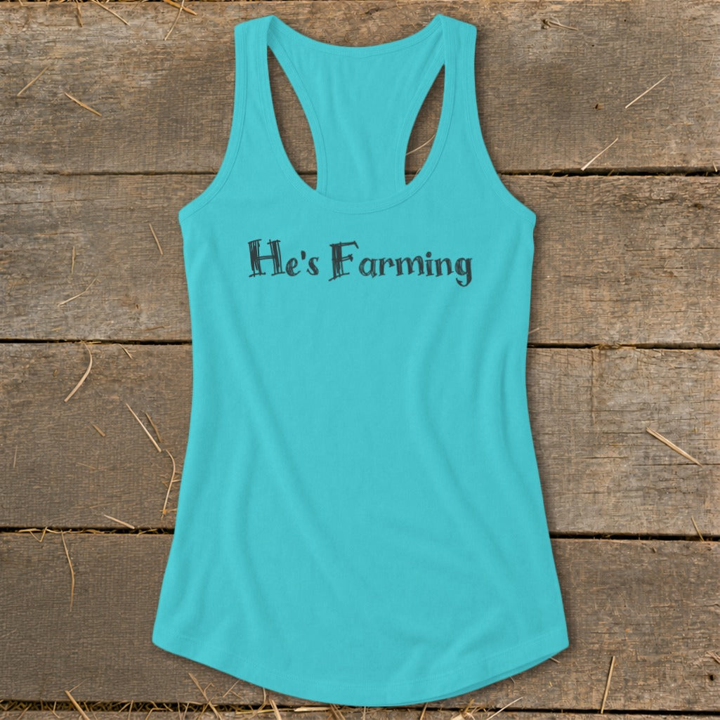 Turquoise tank top with "He's Farming" text on a wooden surface with straw.