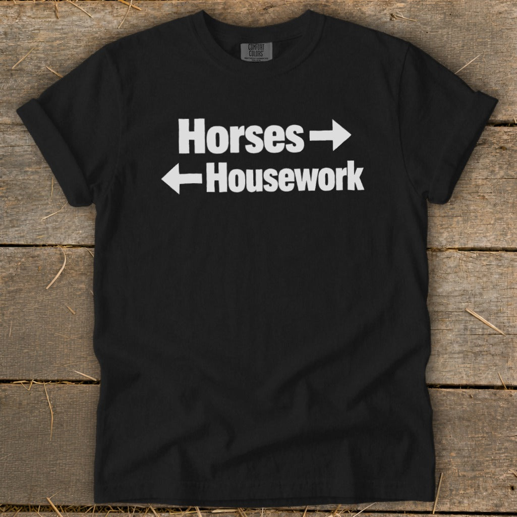 A black crewneck t-shirt with white text that reads Horses with a right arrow and Housework with a left arrow, laid flat on a rustic wooden surface.