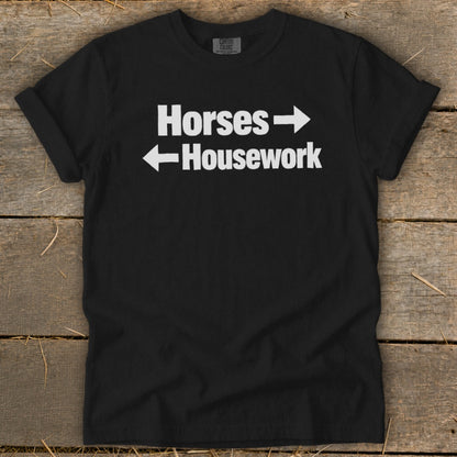 A black crewneck t-shirt with white text that reads Horses with a right arrow and Housework with a left arrow, laid flat on a rustic wooden surface.
