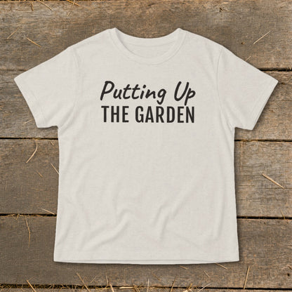 Beige t-shirt with black text "Putting Up THE GARDEN" on a wooden background.