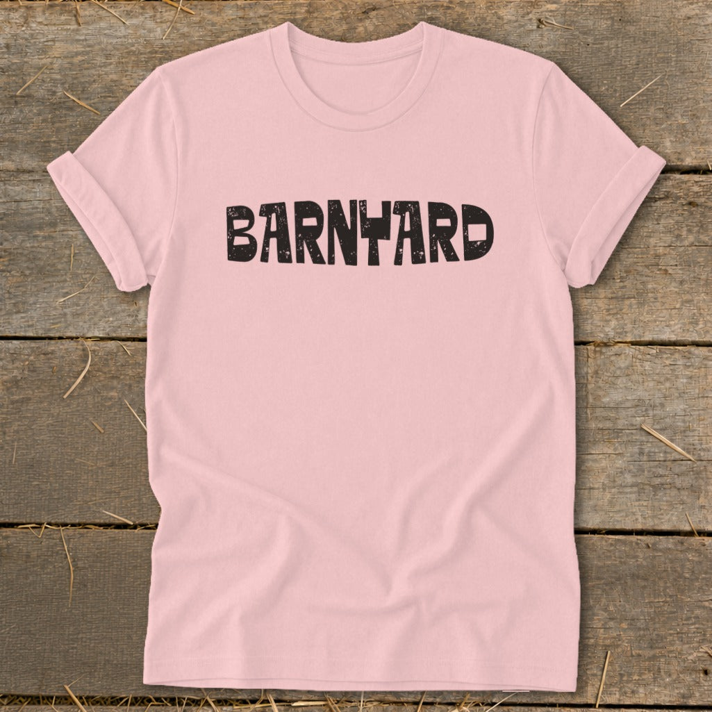 Pink short-sleeve t-shirt laid flat on a rustic wooden background, featuring the distressed black text BARNYARD across the chest.