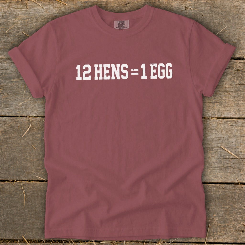 A mauve crewneck t-shirt with distressed white text reading 12 HENS = 1 EGG on a rustic wooden background.