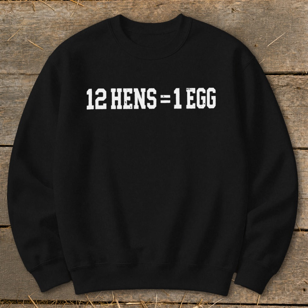 A black crewneck sweatshirt printed with the white text 12 HENS = 1 EGG, laid flat on a rustic wooden surface.
