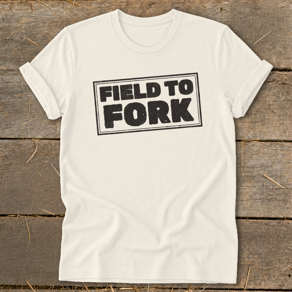 Off-white t-shirt with rolled sleeves laid flat on a wooden surface, featuring black box text reading "FIELD TO FORK".